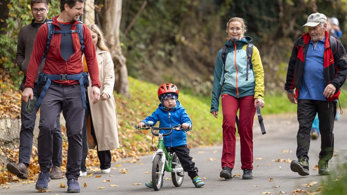 Eine Gruppe von Menschen geht auf einem Weg, während ein Kind auf einem kleinen Fahrrad fährt. Die Umgebung ist herbstlich mit buntem Laub. | © Stadtgemeinde Leoben