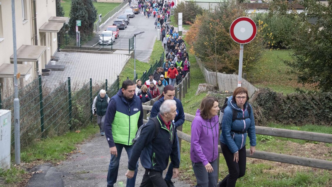 Eine Gruppe von Menschen wandert einen Hügel hinauf, begleitet von einer langen Schlange von Wanderern im Hintergrund. Die Szene zeigt eine grüne Umgebung mit Wohngebäuden und einem klaren Weg. | © Stadtgemeinde Leoben
