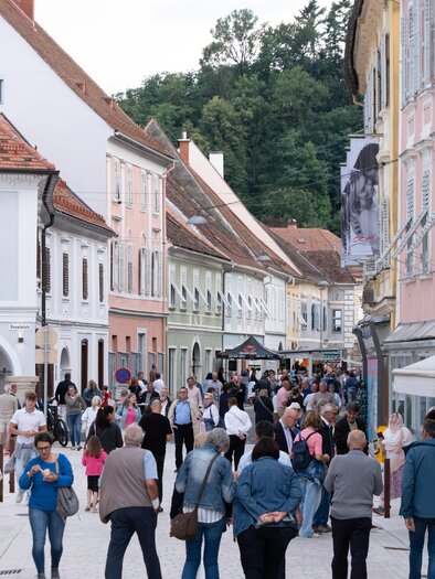 A busy street with many people and colorful buildings. Cafés and shops line the street, surrounded by green trees in the background. | © Flanieren & Radieren