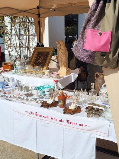 A flea market stall with various antiques and dishes is set up under a sunshade. On the table, there are glass objects, ceramics, and decorative items on display. | © Petra Kirchschlager