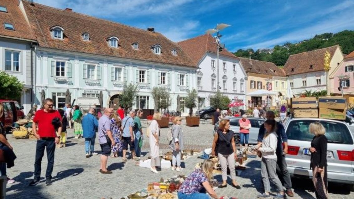Ein belebter Marktplatz mit vielen Menschen, die Stände und Waren betrachten. Im Hintergrund sind historische Gebäuden und ein blauer Himmel zu sehen. | © Oststeiermark Tourismus