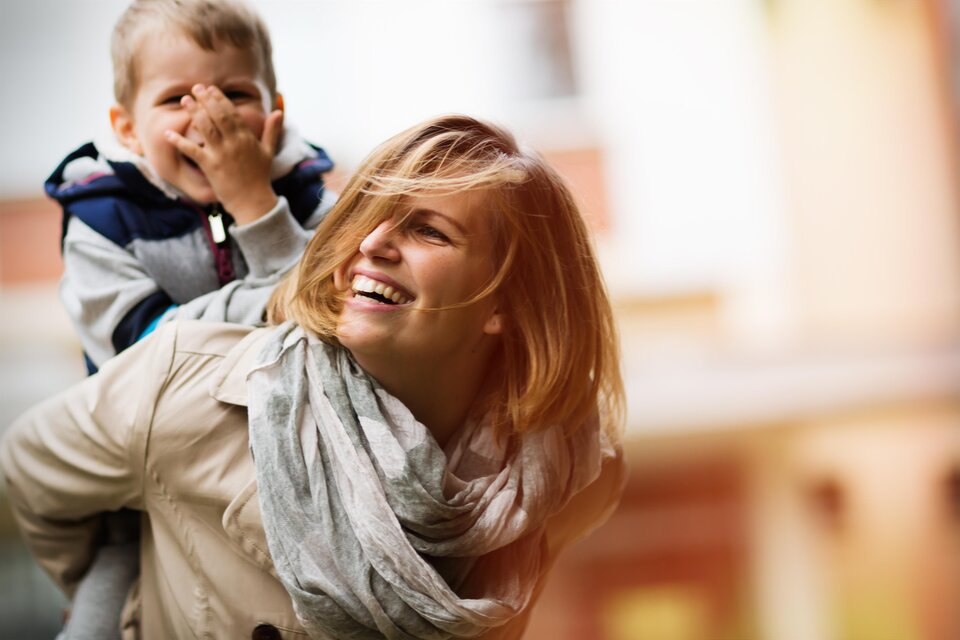 Eine lächelnde Mutter trägt ihr Kind auf dem Rücken. Beide genießen einen fröhlichen Moment im Freien. | © AdobeStock_124019907