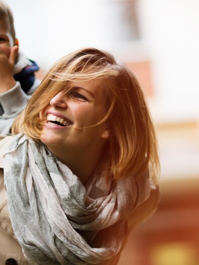 Eine lächelnde Mutter trägt ihr Kind auf dem Rücken. Beide genießen einen fröhlichen Moment im Freien. | © AdobeStock_124019907