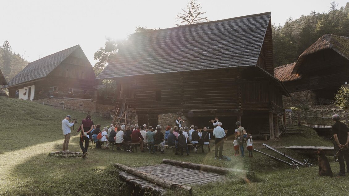 Eine Gruppe von Menschen sitzt im Freien vor einem traditionellen Holzgebäude. Die Szenerie ist von sanften Hügeln und Bäumen umgeben, während die Sonne im Hintergrund scheint. | © Madeleine Marie
