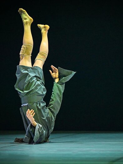 A person is standing upside down on stage and wearing a gray outfit. The background is dark and creates a dramatic atmosphere. | © Oper Graz