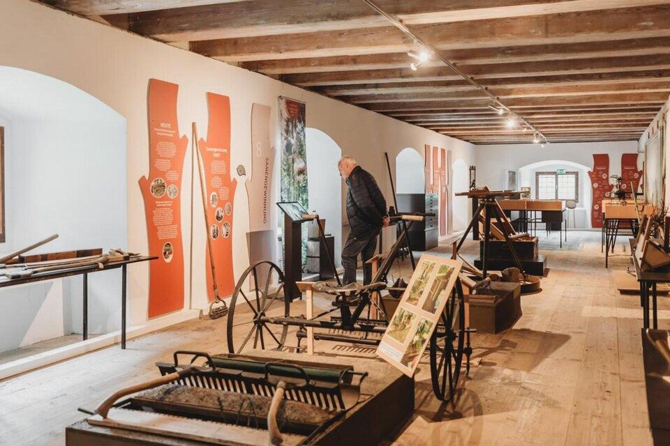 Eine Museumsausstellung mit historischen landwirtschaftlichen Geräten. Im Hintergrund sind informative Tafeln und eine Person zu sehen, die sich die Ausstellung anschaut. | © Forstmuseum Silvanum