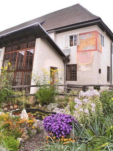Ein charmantes Gebäude mit einem Garten voller bunter Blumen. Im Hintergrund sind sanfte Hügel zu sehen.