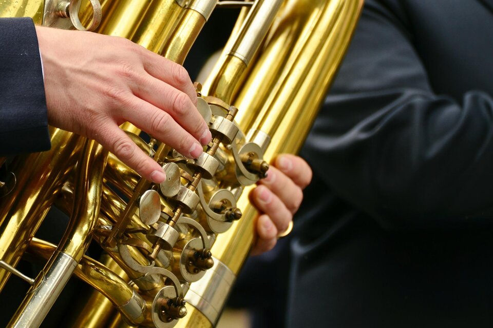 Eine Hand spielt auf einer glänzenden Tuba. Der Hintergrund zeigt einen weiteren Musiker in dunkler Kleidung.