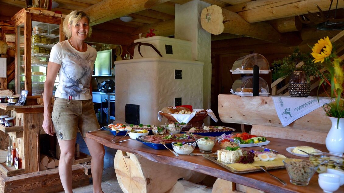 Eine freundliche Frau steht in einer gemütlichen Küche neben einem Tisch voller leckerer Speisen. Der Raum hat rustikale Holzmöbel und eine warme Atmosphäre.