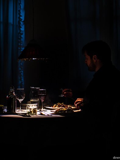 A man is sitting at a table in a dark room, enjoying dinner. The table is set with dishes, glasses, and a bottle of wine.