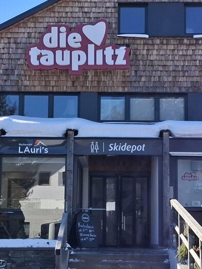 A cozy restaurant with a large window front and a sign with the name "die tauplitz." Snow lies in front of the building, and a few guests can be seen. | © TVB Ausseerland Salzkammergut/Raunigg