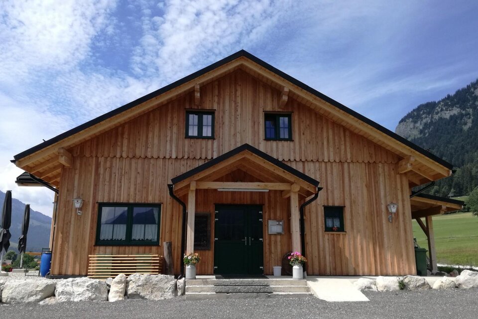 Ein Holzhaus mit einem attraktiven Eingang und großen Fenstern. Im Hintergrund sind grüne Wiesen und Berge sichtbar. | © Stoffis Hütten