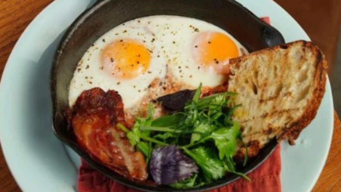 Ein herzhaftes Frühstücksgericht in einer kleinen Pfanne. Es besteht aus zwei Spiegeleiern, gebratenem Speck, einer Scheibe Brot und frischem Gemüse. | © Zum fliegenden Honigtopf