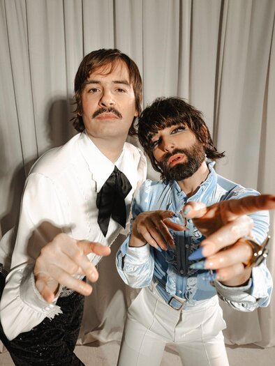 Martin Zerza als Herr Martin - in weißem Hemd, mit schwarzer Schleife und scharzer Hose - und Tom Neuwirth als Frau Martin - in hellblauem Rüschenhemd und weißer Hose, mit langen Fingernägeln, Schminke und Vollbart - präsentieren sich vor einem beigen Bühnenvorhang. | © Oststeiermark Tourismus