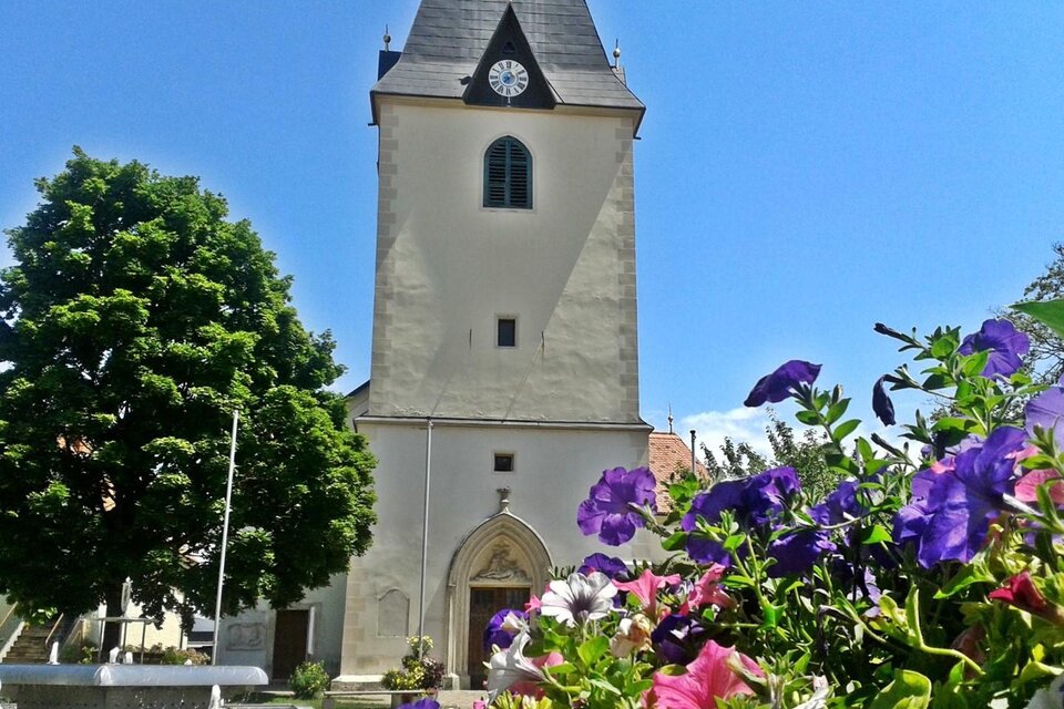 Eine alte Kirche mit einem hohen Turm und einem Ziffernblatt. Bunte Blumen umrahmen den Vordergrund der Szene. | © Pfarre Gamlitz