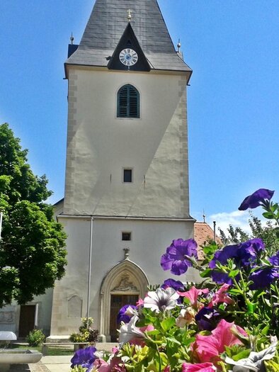 Eine alte Kirche mit einem hohen Turm und einem Ziffernblatt. Bunte Blumen umrahmen den Vordergrund der Szene. | © Pfarre Gamlitz