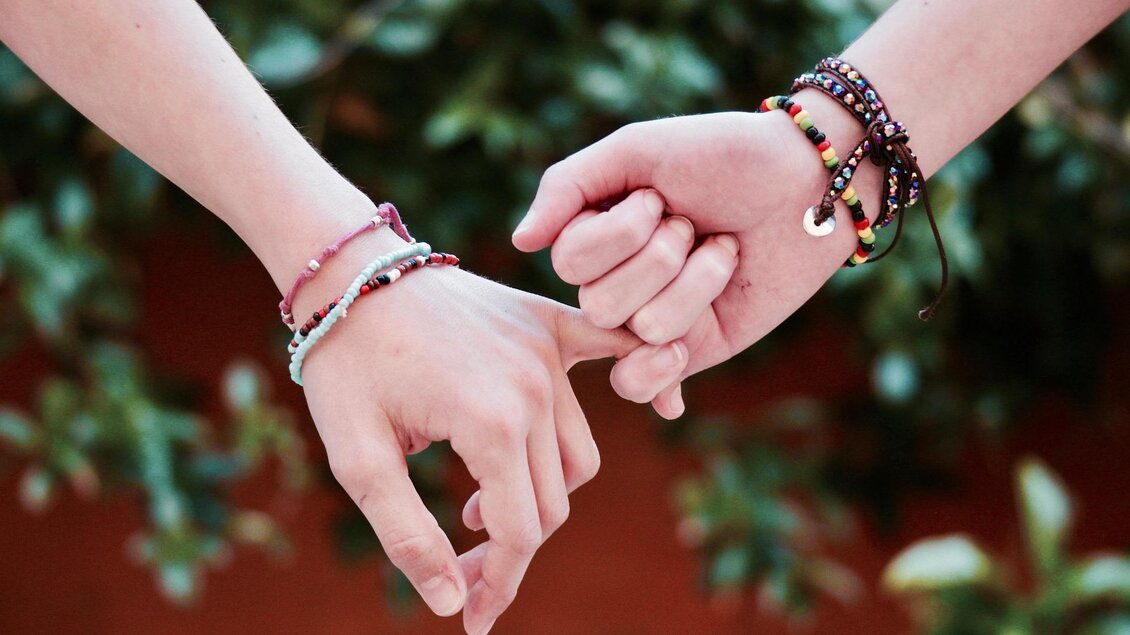 Zwei Hände halten sich gegenseitig an den Fingern. Sie sind mit bunten Armbändern geschmückt und umgeben von einer grünen Pflanzenkulisse. | © Pixabay