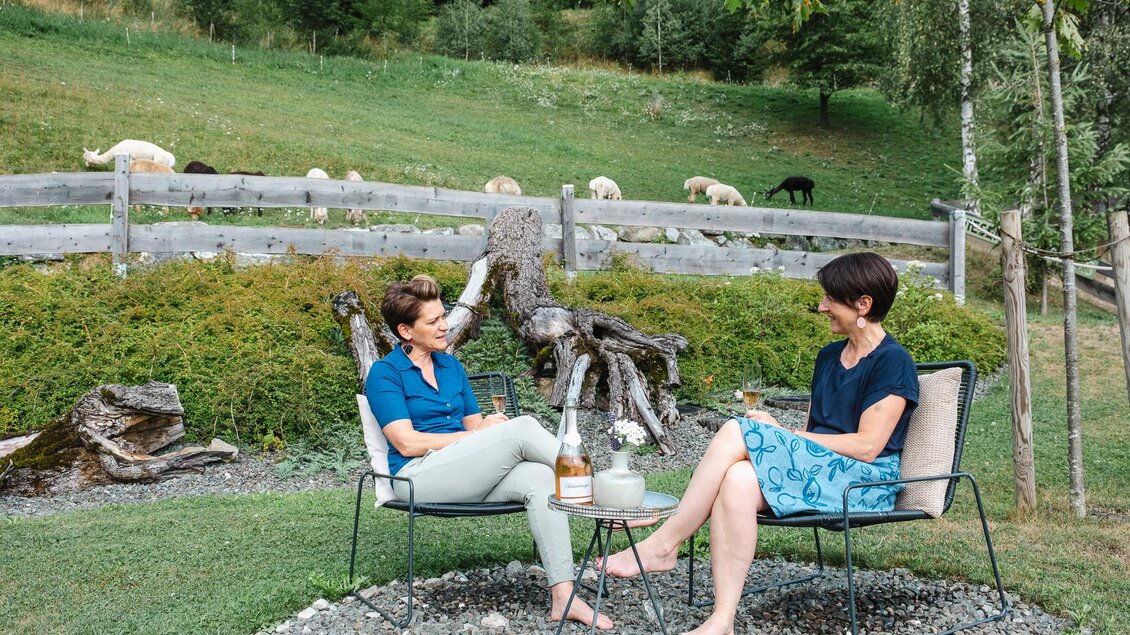 Zwei Frauen sitzen entspannt im Freien und unterhalten sich. Im Hintergrund sieht man eine Wiese mit Schafen und Bäumen. | © Wieserhof_Freundinnenzeit