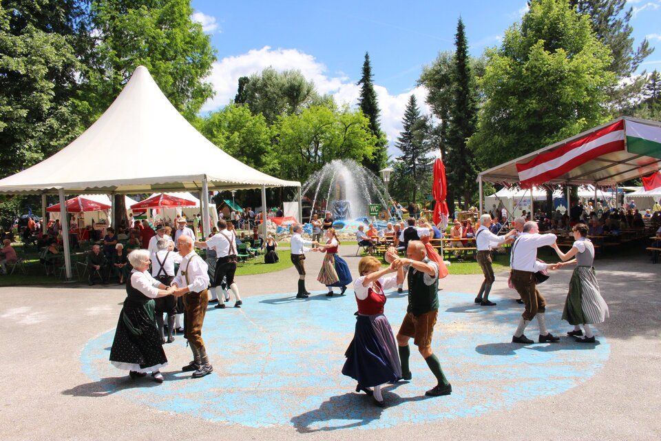 Eine Gruppe von Menschen tanzt fröhlich in traditionellen Trachten in einem Park. Im Hintergrund sind Zelte und Bäume zu sehen. | © Kulturreferat Bärnbach