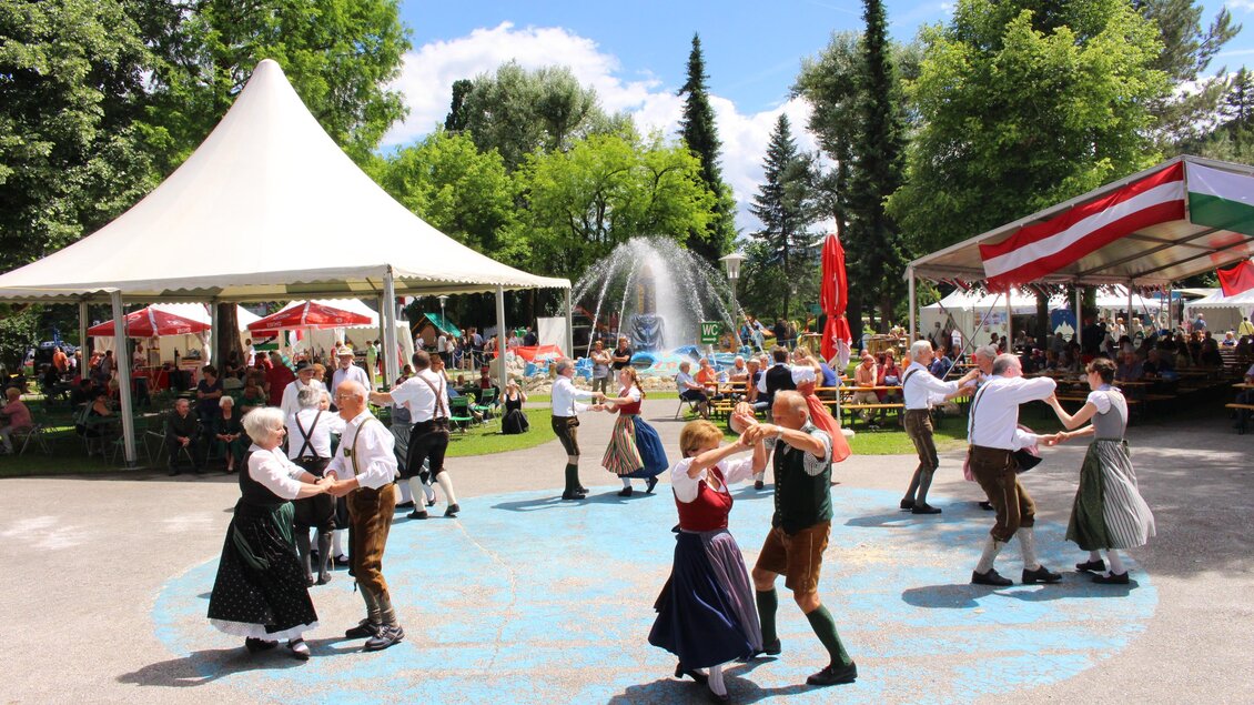 Eine Gruppe von Menschen tanzt fröhlich in traditionellen Trachten in einem Park. Im Hintergrund sind Zelte und Bäume zu sehen. | © Kulturreferat Bärnbach