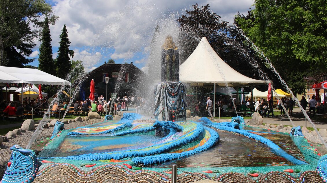 Ein brunnenspielfülltes Becken mit bunten Mosaiken und sprudelndem Wasser. Im Hintergrund sind Zelte und Menschen zu sehen, die den Tag genießen. | © Kulturreferat Bärnbach