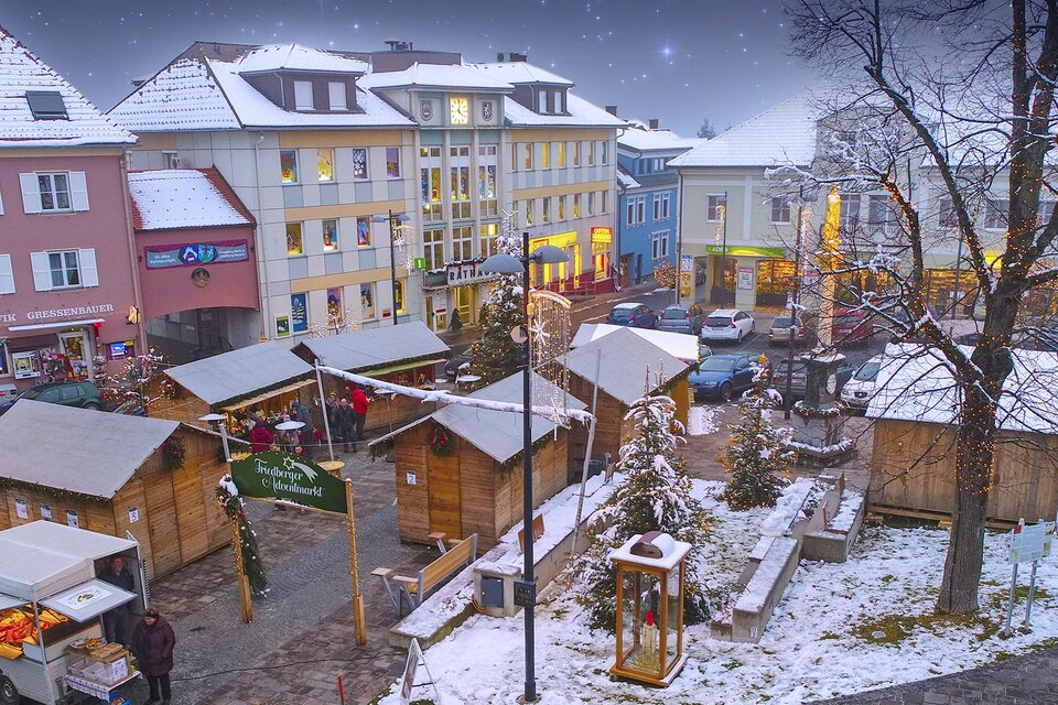 Ein malerischer Weihnachtsmarkt in einer Stadt mit schneebedeckten Ständen. Die Umgebung ist festlich beleuchtet und es liegt Schnee auf den Dächern. | © Stadtgemeinde Friedberg