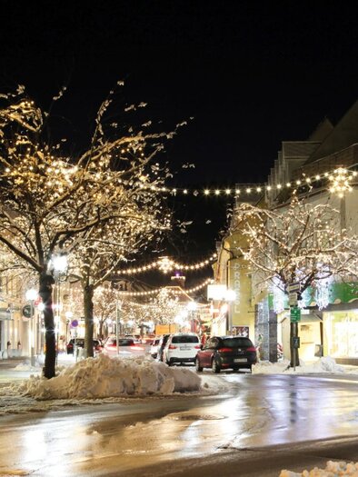 A charming street with festive lights and snow-covered trees at night. The buildings are brightly lit, creating a cozy atmosphere.