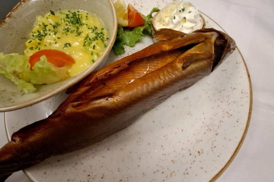 Ein geräucherter Fisch auf einem Teller, begleitet von einem Salat und einer cremigen Beilage. Neben dem Fisch liegt ein Stück Zitrone und ein Klecks Sauerrahm. | © Kainzinger