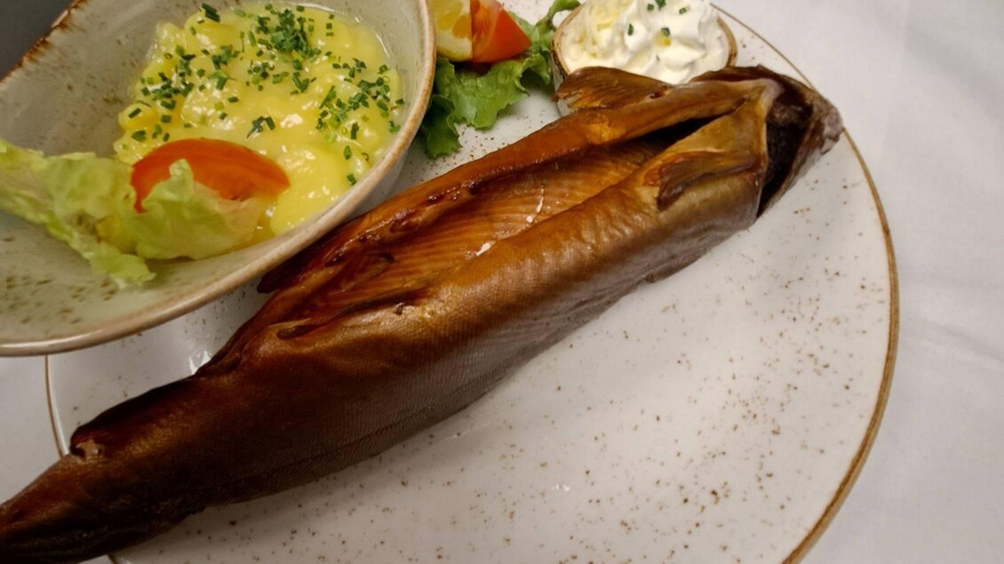 Ein geräucherter Fisch auf einem Teller, begleitet von einem Salat und einer cremigen Beilage. Neben dem Fisch liegt ein Stück Zitrone und ein Klecks Sauerrahm. | © Kainzinger