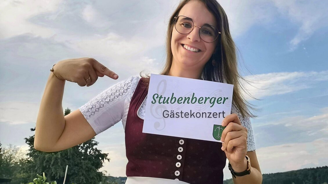 Eine fröhliche Frau in traditioneller Tracht hält ein Schild mit der Aufschrift "Stubenberger Gästekonzert". Im Hintergrund sind Wiesen und ein blauer Himmel zu sehen. | © Lisa Lenz_Oststeiermark
