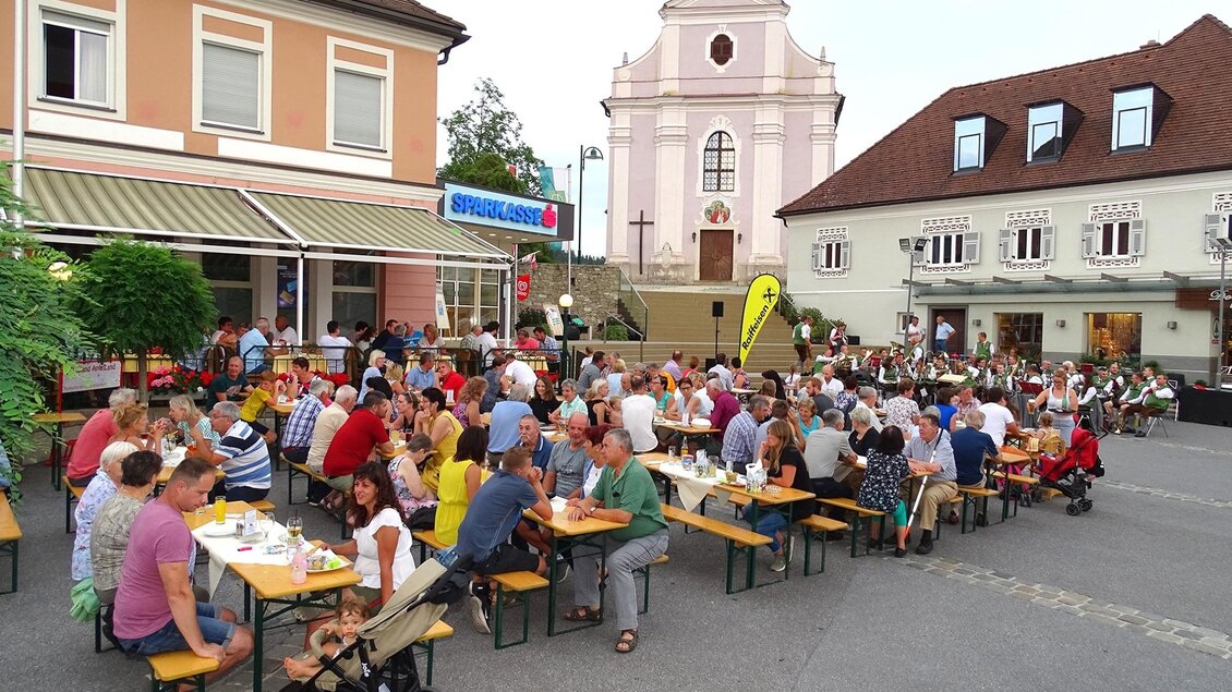 Eine belebte Straßenszene mit vielen Menschen, die an langen Tischen im Freien essen. Im Hintergrund ist eine Kirche und mehrere Gebäude zu sehen. | © Gästekonzert_Oststeiermark