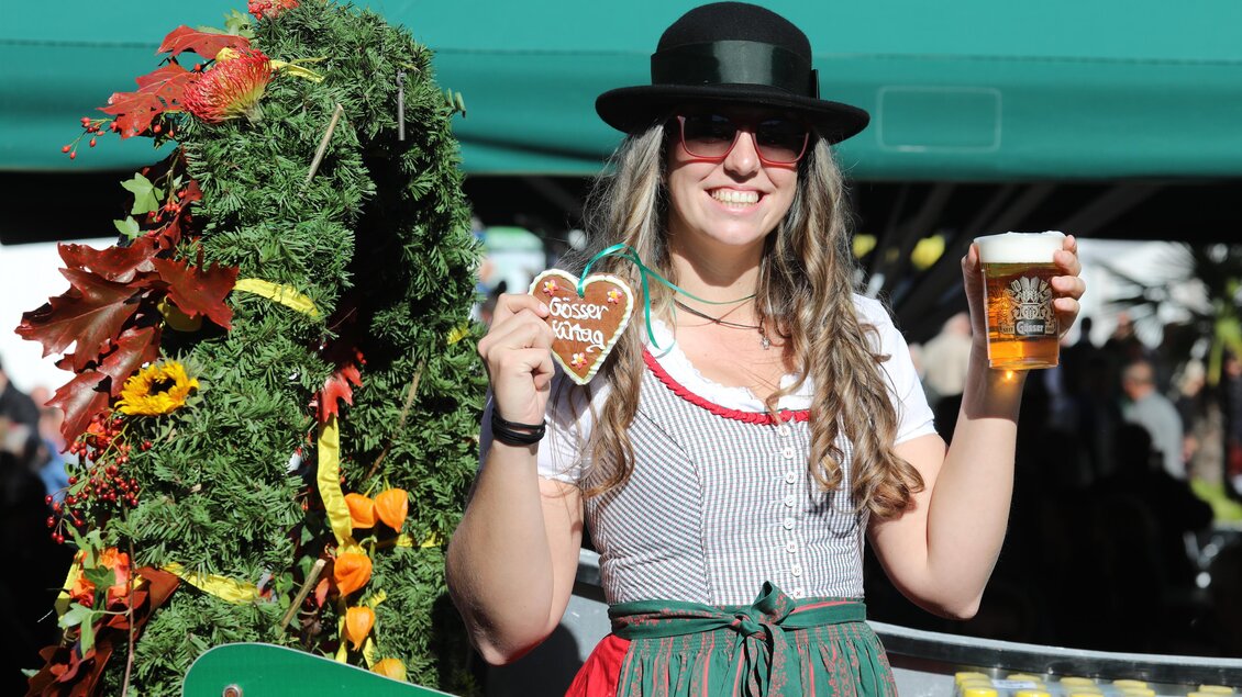 Eine fröhliche Frau trägt ein traditionelles Outfit und hält ein Bier sowie ein Lebkuchenherz in der Hand. Im Hintergrund sind festliche Dekorationen und Getränke sichtbar. | © Foto Freisinger