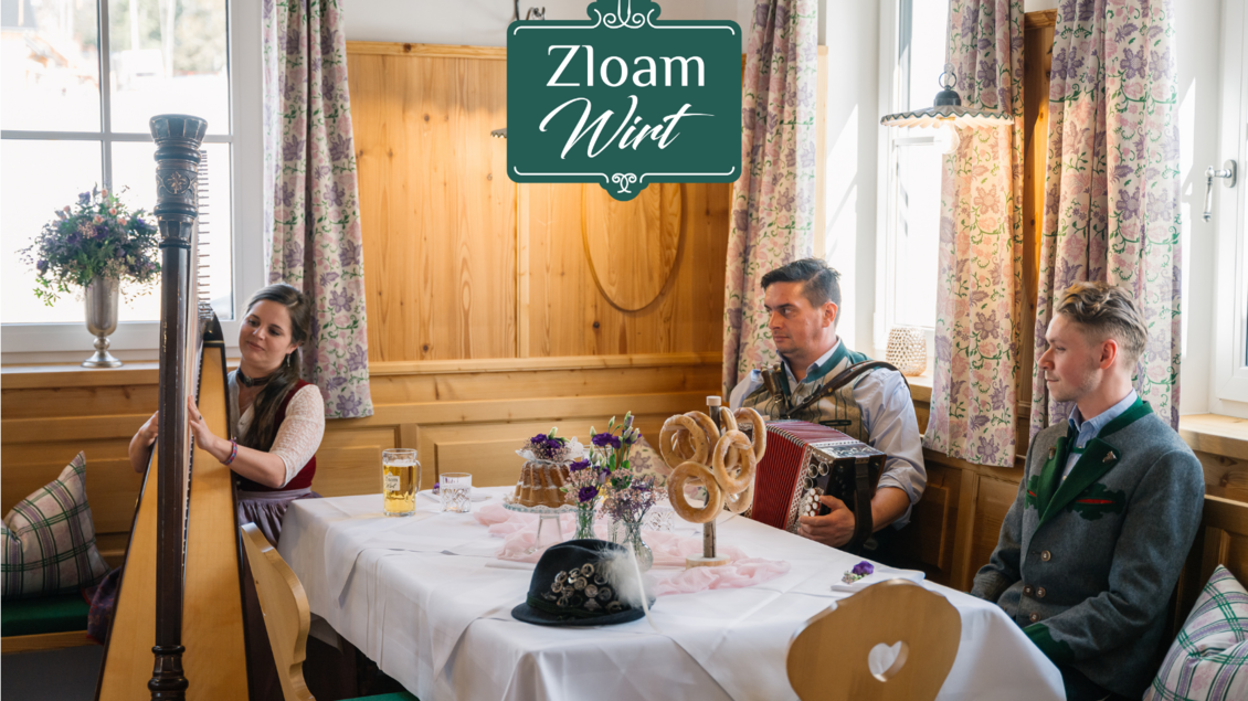 Eine gemütliche Stube mit traditioneller Einrichtung. Musiker spielen auf einer Harfe und einem Akkordeon, während eine bierhaltige Getränk auf dem Tisch steht. | © www.zloam.at