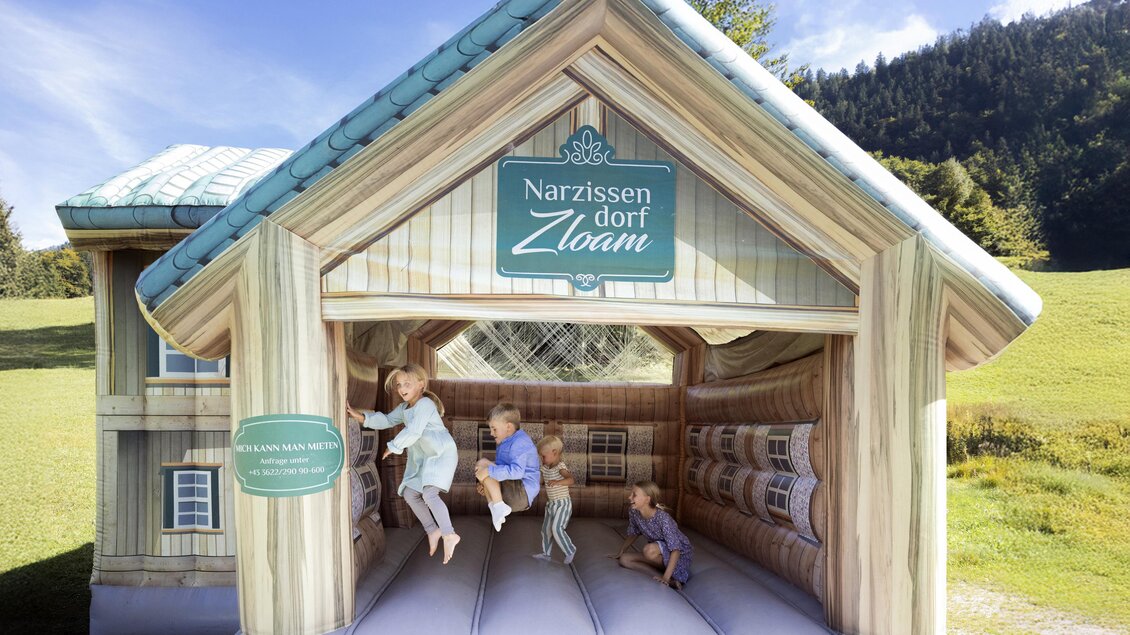 Ein aufblasbares Spielhaus mit dem Namen "Narzissen Dorf Zloam". Kinder spielen und hüpfen darin auf grüner Wiese mit Bergen im Hintergrund. | © www.zloam.at