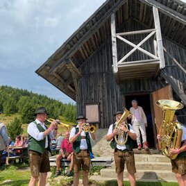Eine Blaskapelle spielt vor einer Holzschutzhütte in der Natur. Im Hintergrund stehen Menschen und genießen die Musik. | © Erlebnisregion Murtal