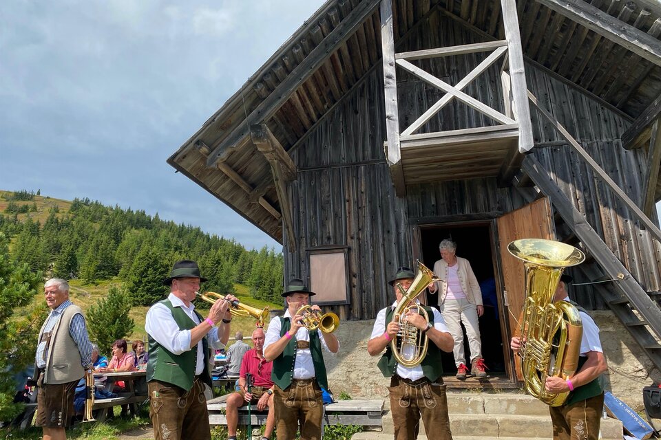 Eine Blaskapelle spielt vor einer Holzschutzhütte in der Natur. Im Hintergrund stehen Menschen und genießen die Musik. | © Erlebnisregion Murtal