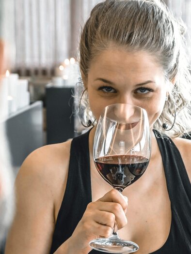 <p>Eine Frau hält ein Weinglas und schaut lächelnd über den Rand. Im Hintergrund ist eine gemütliche Restaurantatmosphäre im Vulkanlandhotel Legenstein in Bairisch Kölldorf zu sehen.</p> | © Vulkanlandhotel Legenstein