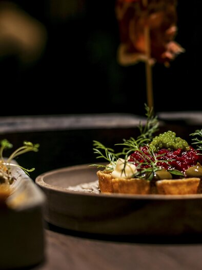 An elegant dish on a plate, decorated with fresh herbs and a colorful garnish. In the background, more appetizing foods can be seen. | © jamst_winzertisch 220324_42
