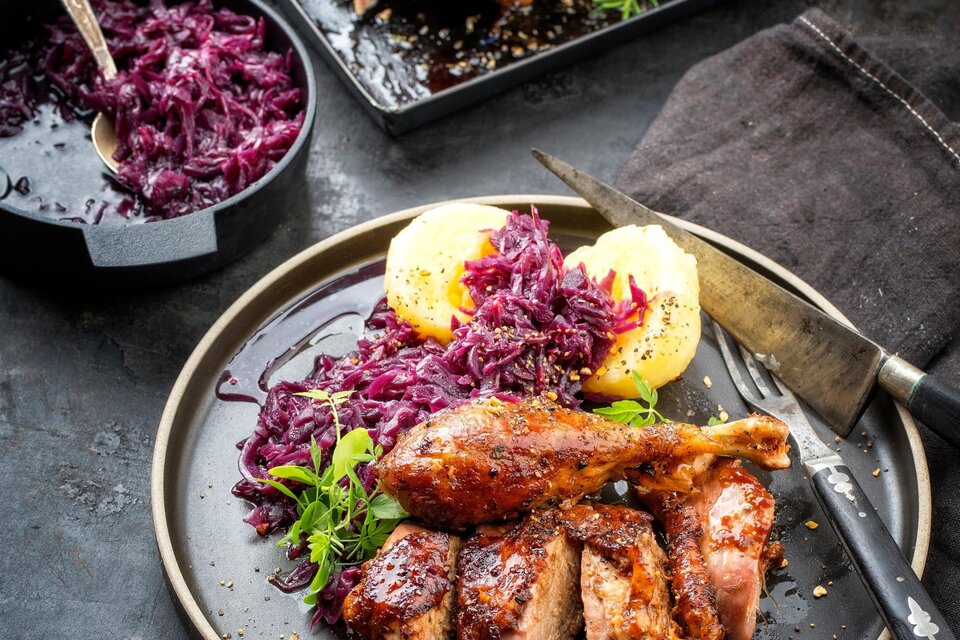 Ein Teller mit saftigem Fleisch und Rotkohl, garniert mit Kartoffeln. Im Hintergrund ist ein gebratenes Fleischstück zu sehen. | © TV Südsteiermark AdobeStock_239581696