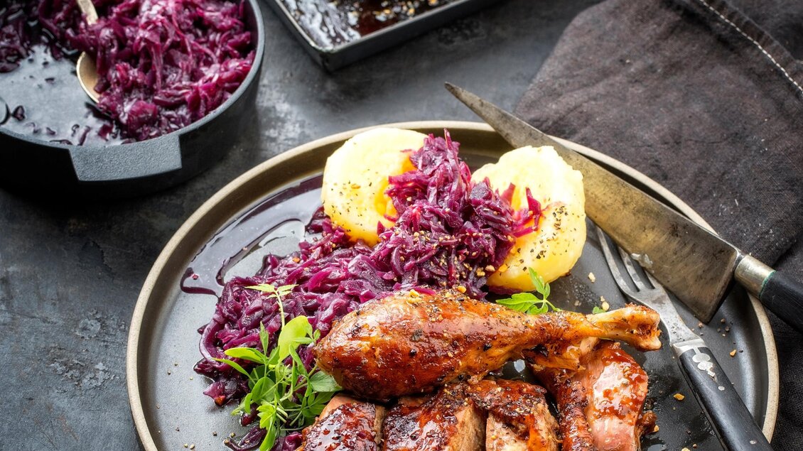 Ein Teller mit saftigem Fleisch und Rotkohl, garniert mit Kartoffeln. Im Hintergrund ist ein gebratenes Fleischstück zu sehen. | © TV Südsteiermark AdobeStock_239581696