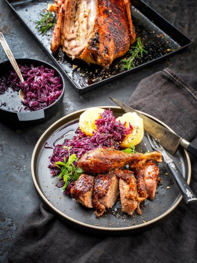 A plate with juicy meat and red cabbage, garnished with potatoes. In the background, a piece of roast meat is visible. | © TV Südsteiermark AdobeStock_239581696