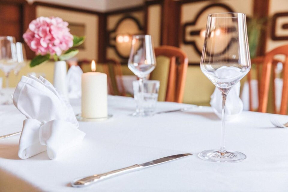 Ein schön gedeckter Tisch in einem Restaurant mit einer weißen Tischdecke. Im Vordergrund stehen eine Glaskaraffe, ein Weinglas und eine brennende Kerze. | © Landgasthof Wratschko