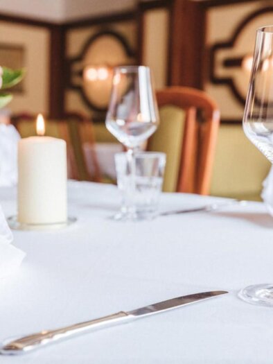 Ein schön gedeckter Tisch in einem Restaurant mit einer weißen Tischdecke. Im Vordergrund stehen eine Glaskaraffe, ein Weinglas und eine brennende Kerze. | © Landgasthof Wratschko