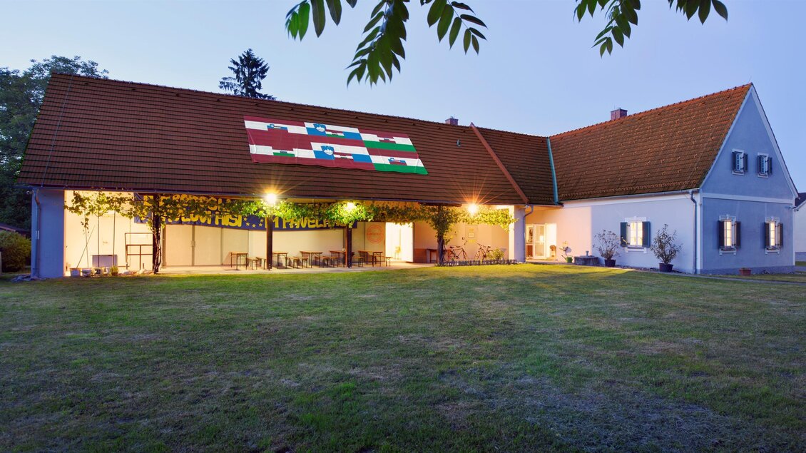 Ein schönes Haus mit einem großen Garten, das in der Abenddämmerung beleuchtet ist. Auf dem Dach ist eine bunte Flagge zu sehen. | © Pavelhaus