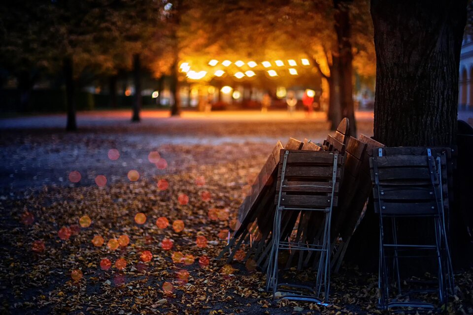 Ein herbstlicher Biergarten in der Dämmerung, mit zusammengeklappten Holztischen und -stühlen unter Bäumen, deren Laub den Boden bedeckt. Im Hintergrund leuchten warme Lichter in der Ferne und erzeugen eine gemütliche Atmosphäre. | © Pixabay