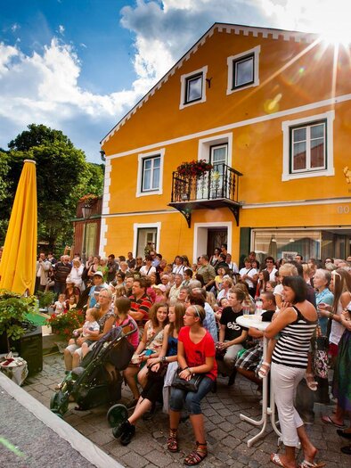 A lively crowd gathers on a street in front of a yellow building. A woman walks elegantly across the sidewalk while the spectators watch attentively.