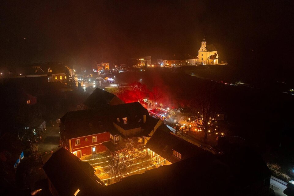 Eine stimmungsvolle Nachtaufnahme eines kleinen Dorfes mit beleuchteten Gebäuden und einer Kirche im Hintergrund. Der Himmel ist dunkel, und es gibt viele Lichter, die eine festliche Atmosphäre schaffen. | © St. Johanner Gassl-Advent