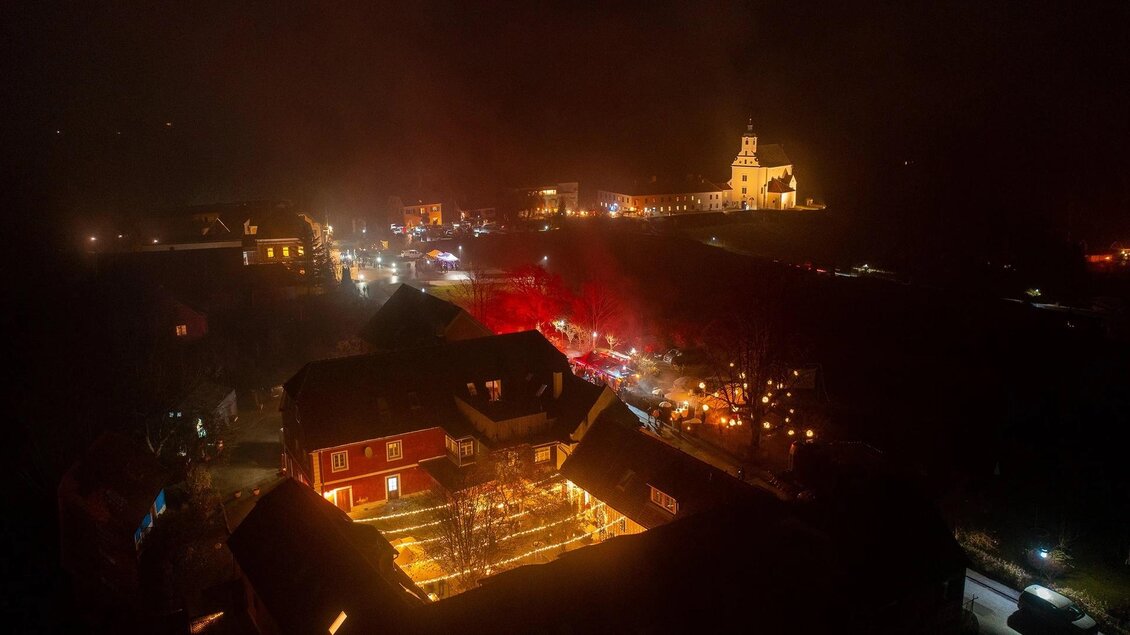 Eine stimmungsvolle Nachtaufnahme eines kleinen Dorfes mit beleuchteten Gebäuden und einer Kirche im Hintergrund. Der Himmel ist dunkel, und es gibt viele Lichter, die eine festliche Atmosphäre schaffen. | © St. Johanner Gassl-Advent