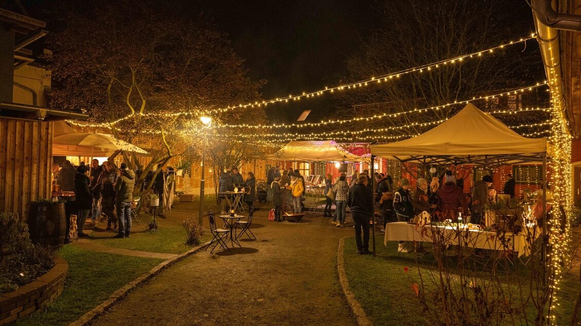 Ein festlicher Abendmarkt mit vielen Menschen unter Lichtern. Stände bieten verschiedene Waren an und die Atmosphäre ist lebhaft und einladend. | © Gassl Advent
