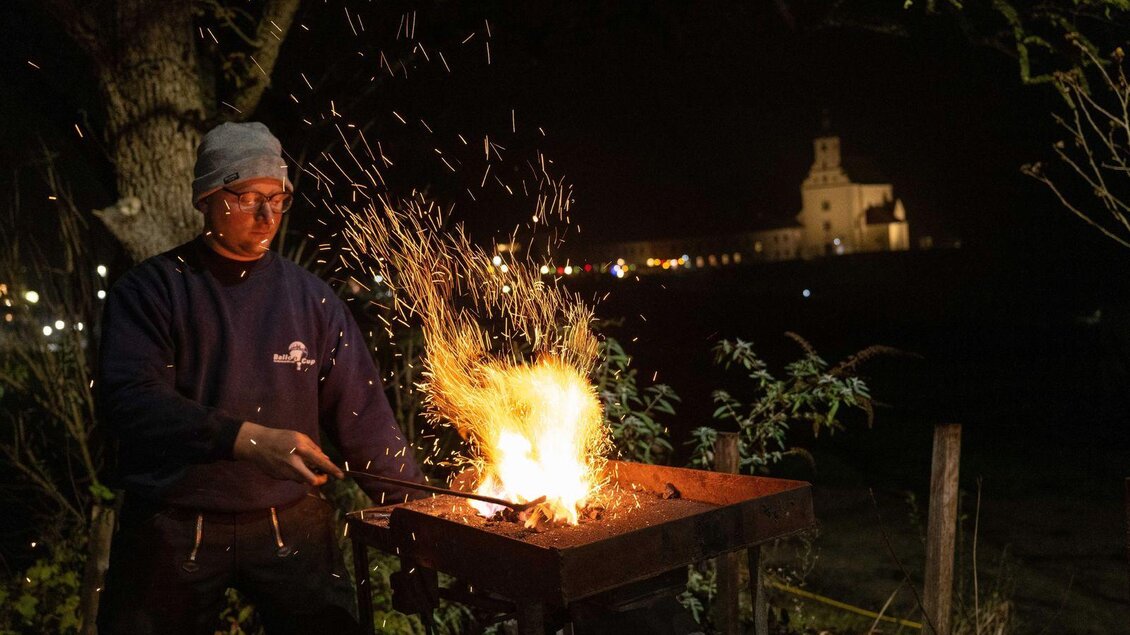Ein Schmied arbeitet an einer Schmiede mit leuchtenden Funken. Im Hintergrund ist ein beleuchtetes Gebäude bei Nacht zu sehen. | © Gassladvent